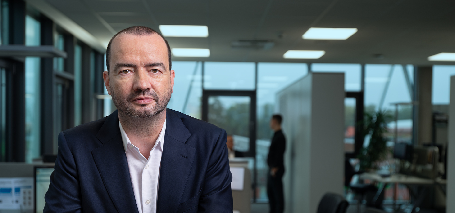 Porträt von Ronny Stagen in einem modernen Büro mit Büromöbeln und weiteren Personen im Hintergrund.
